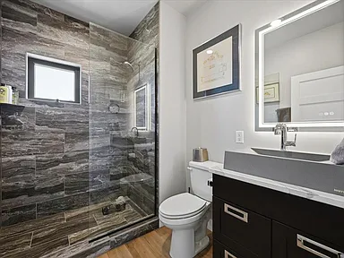 A stone-tiled walk-in shower stands beside a toilet and dark vanity with an arched mirror in this bathroom.