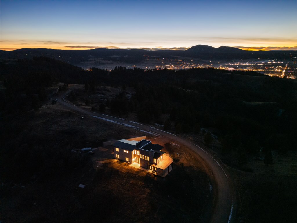 At night, the house is lit from within on its ridge, with the wide grid of town lights filling the valley beyond.