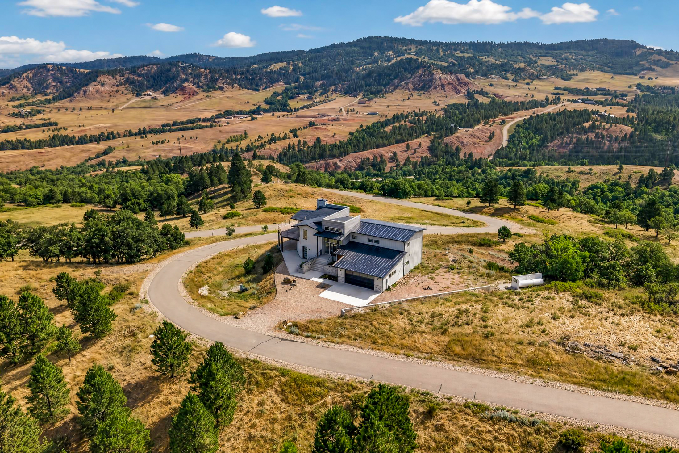 From above, the house sits before a backdrop of red badlands-style ridgelines and forested hills.