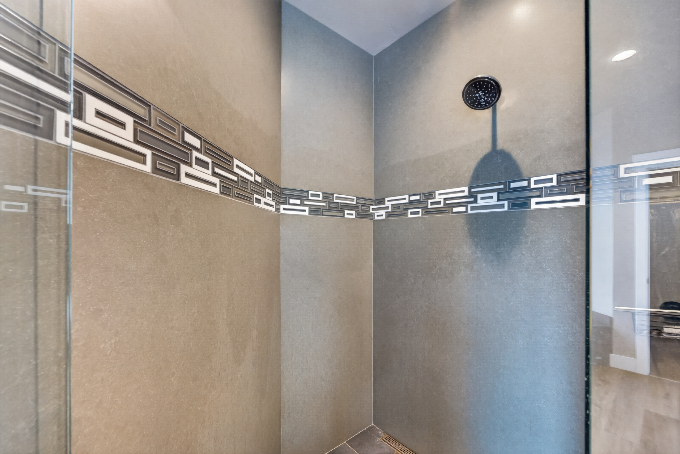 A glass-enclosed shower corner shows a rain showerhead and a decorative mosaic tile band running along the walls.